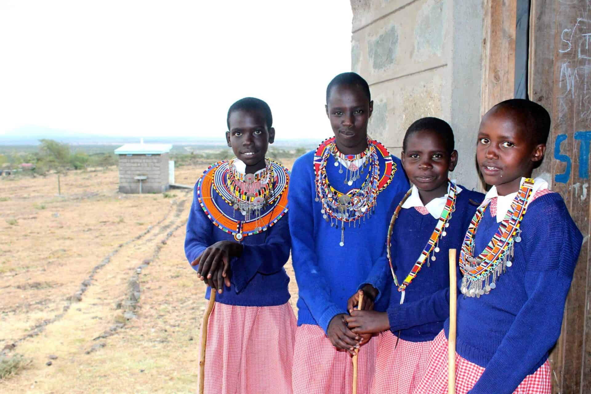 Four students stand outside the Endonyio Sidai Primary School. Each girl is wearing traditional Maasai beaded necklaces over their school uniforms of long-sleeved blue sweaters over white collared blouses and red and white gingham skirts. Behind them is a well house.