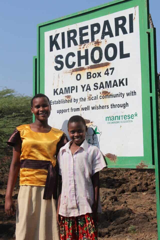 Primary school students stand outside the Kirepari School on Kokwa Island in rural Kenya. The girl on the left is wearing a yellow satin shirt with a brown sash and a light-colored skirt. The girl on the right is shorter and wearing a light-colored collared shirt and a red skirt. They are both smiling.