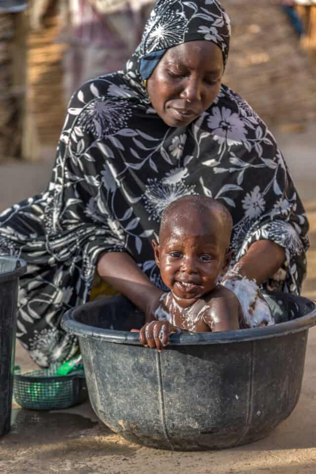 A woman washes her child in a basin with clean water. She is wearing a black dress and head wrap with white floral print. Her baby is smiling at the camera.