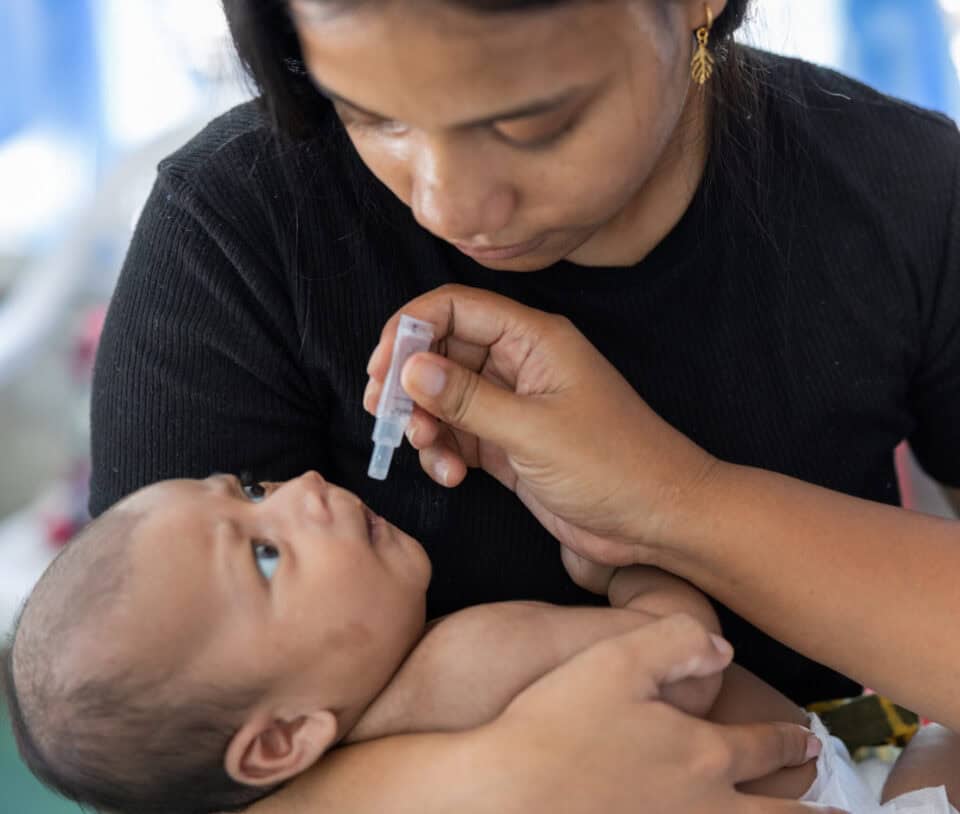 An infant receives a vaccination while being held. A healthcare worker administers a drop in its mouth while the child looks up at the mother.