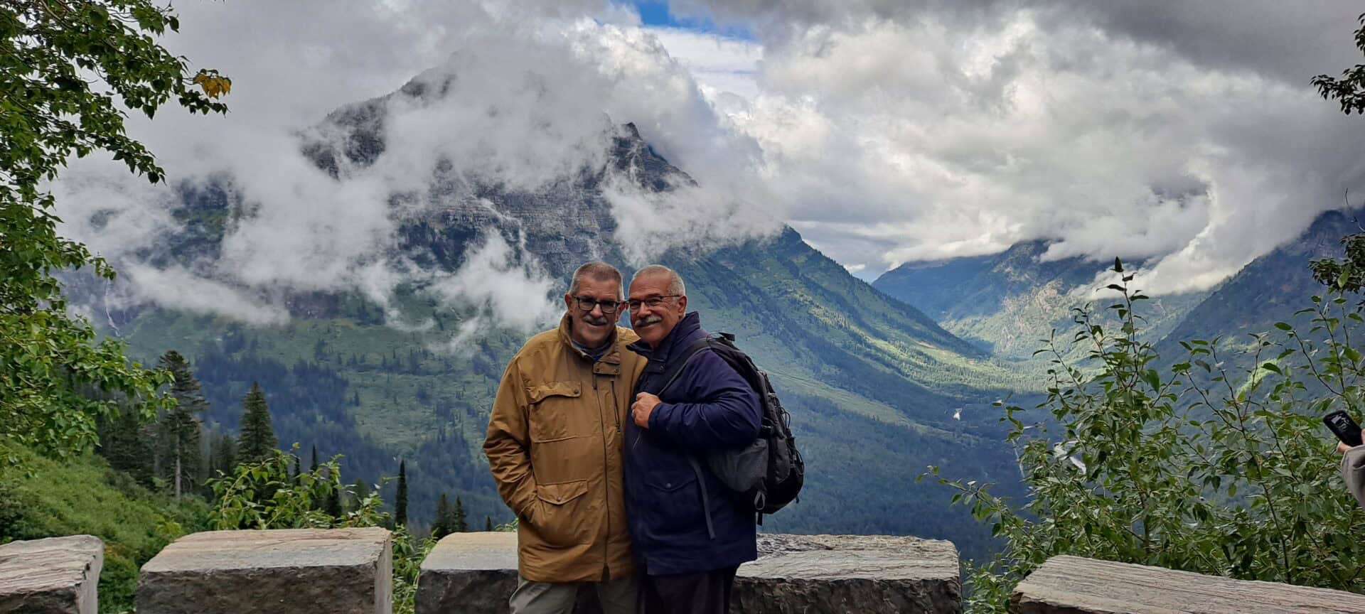 Grandpa Bobby and Grandpa Scott smile in front of a sweeping landscape. Clouds hover around a massive mountain behind them.