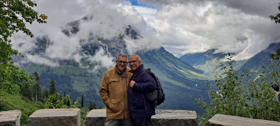 Grandpa Bobby and Grandpa Scott smile in front of a sweeping landscape. Clouds hover around a massive mountain behind them.