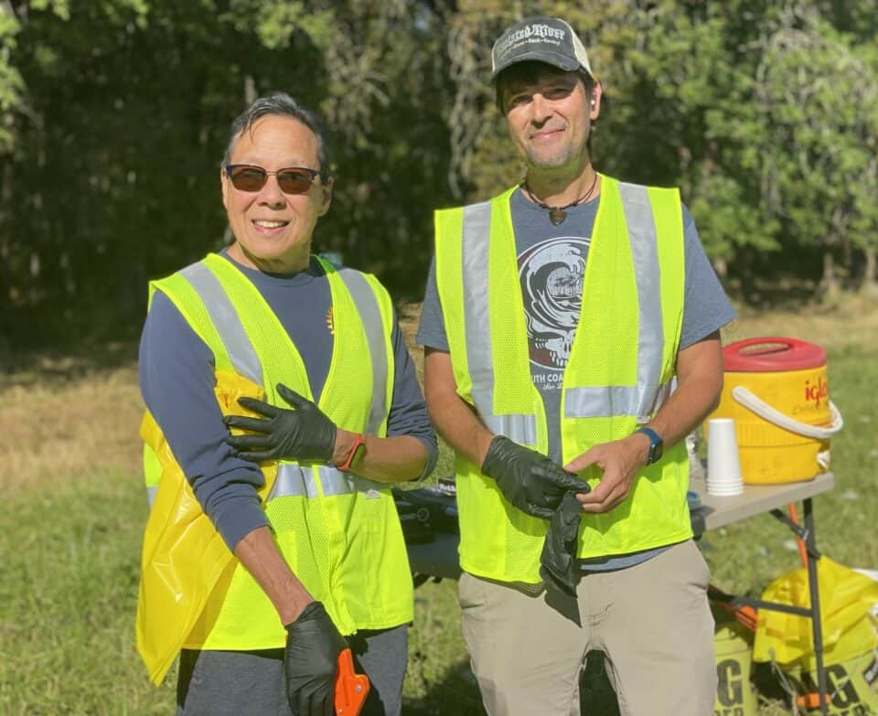 Two men smile for the camera while picking up trash at the Amazon Creek. They are wearing reflective vests and rubber gloves.