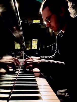 Pianist Andrei Andreev is illuminated from the side in a low-lit room. He is playing intently, leaning over the piano and the keys stretch toward the foreground of the photo.