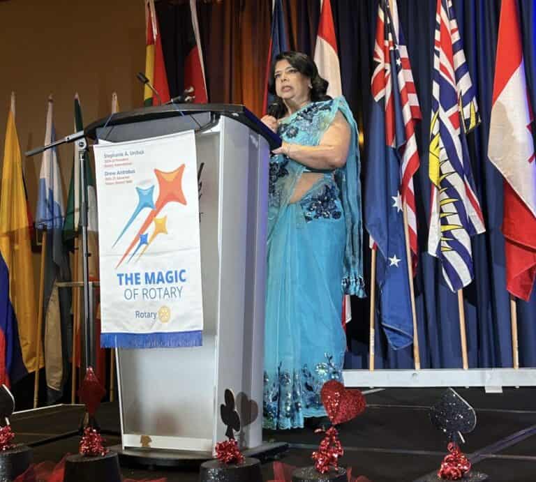 Rumana Monzur speaks at the 2025 District Conference. She is on stage behind a podium with the Magic of Rotary flag and behind her are flags of the world. She is wearing a beautiful blue saree and holding the microphone.