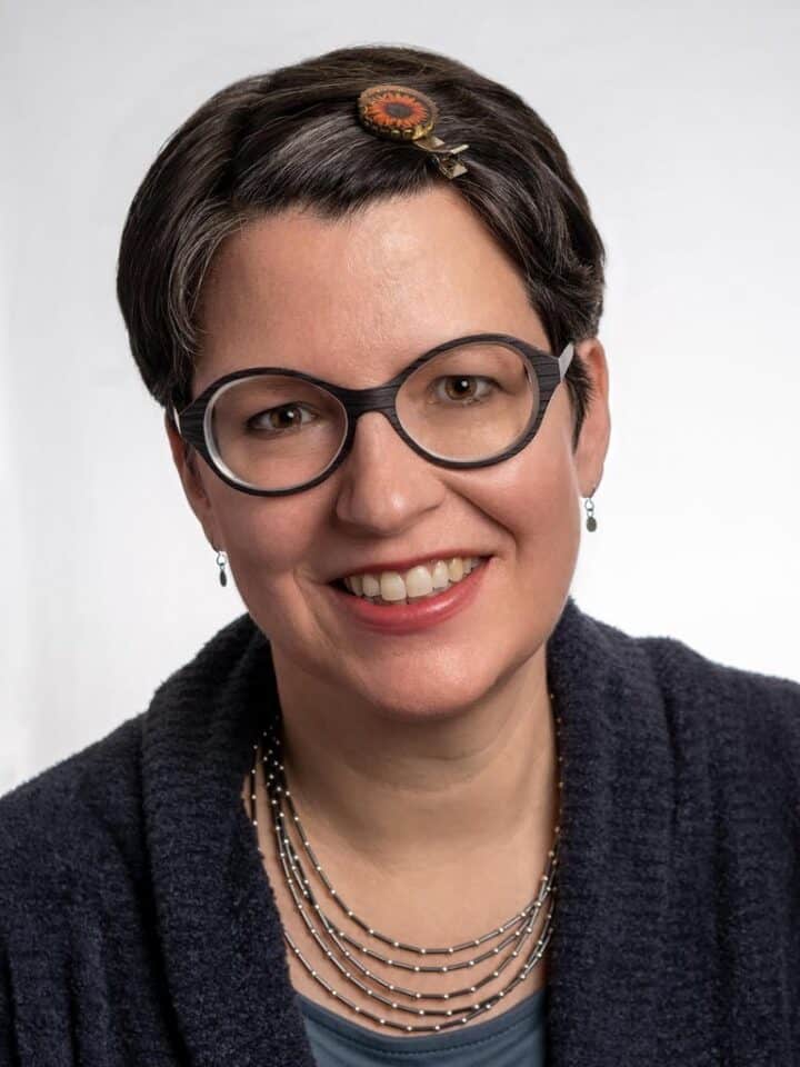 Kate Silver smiles at the camera. She has short dark hiar, dark framed glasses and is wearing a black blouse with layered necklaces and small earrings.