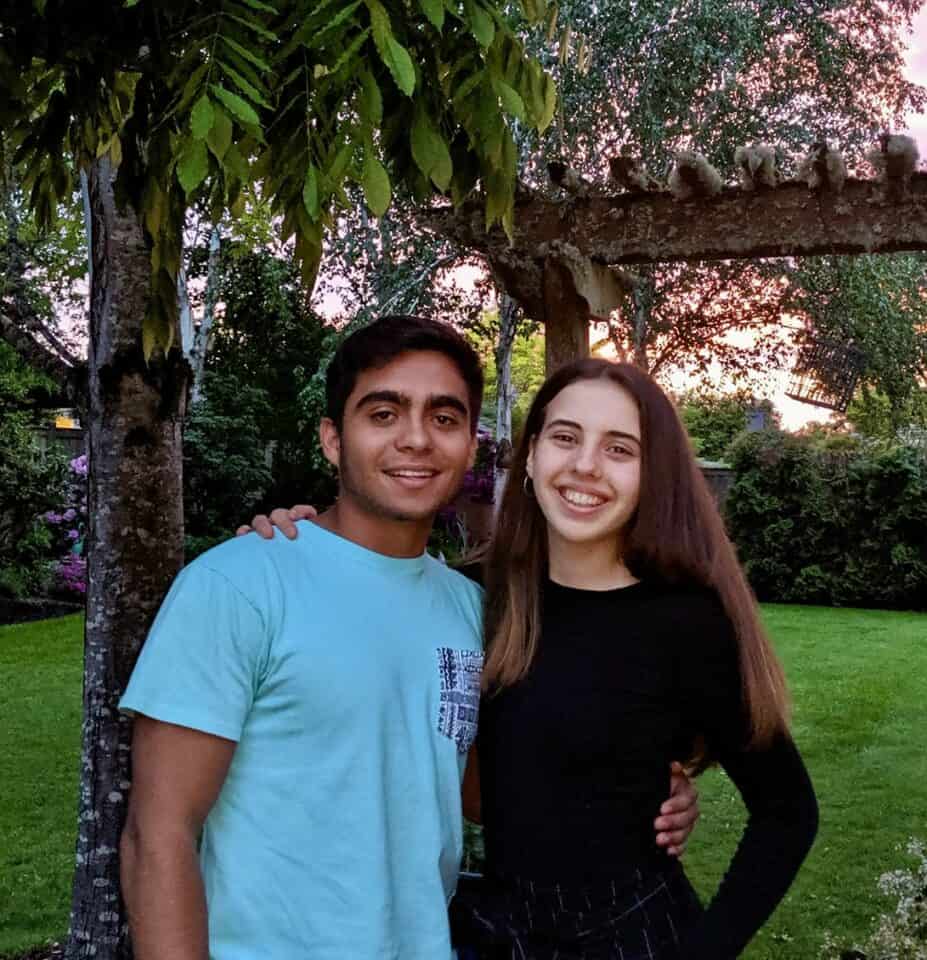 Two Rotary Youth Exchange students smile for the camera with their arms around each other. They are standing outside at sunset in front of a green lawn. He is wearing a blue tshirt and she is wearing a long-sleeved black shirt.