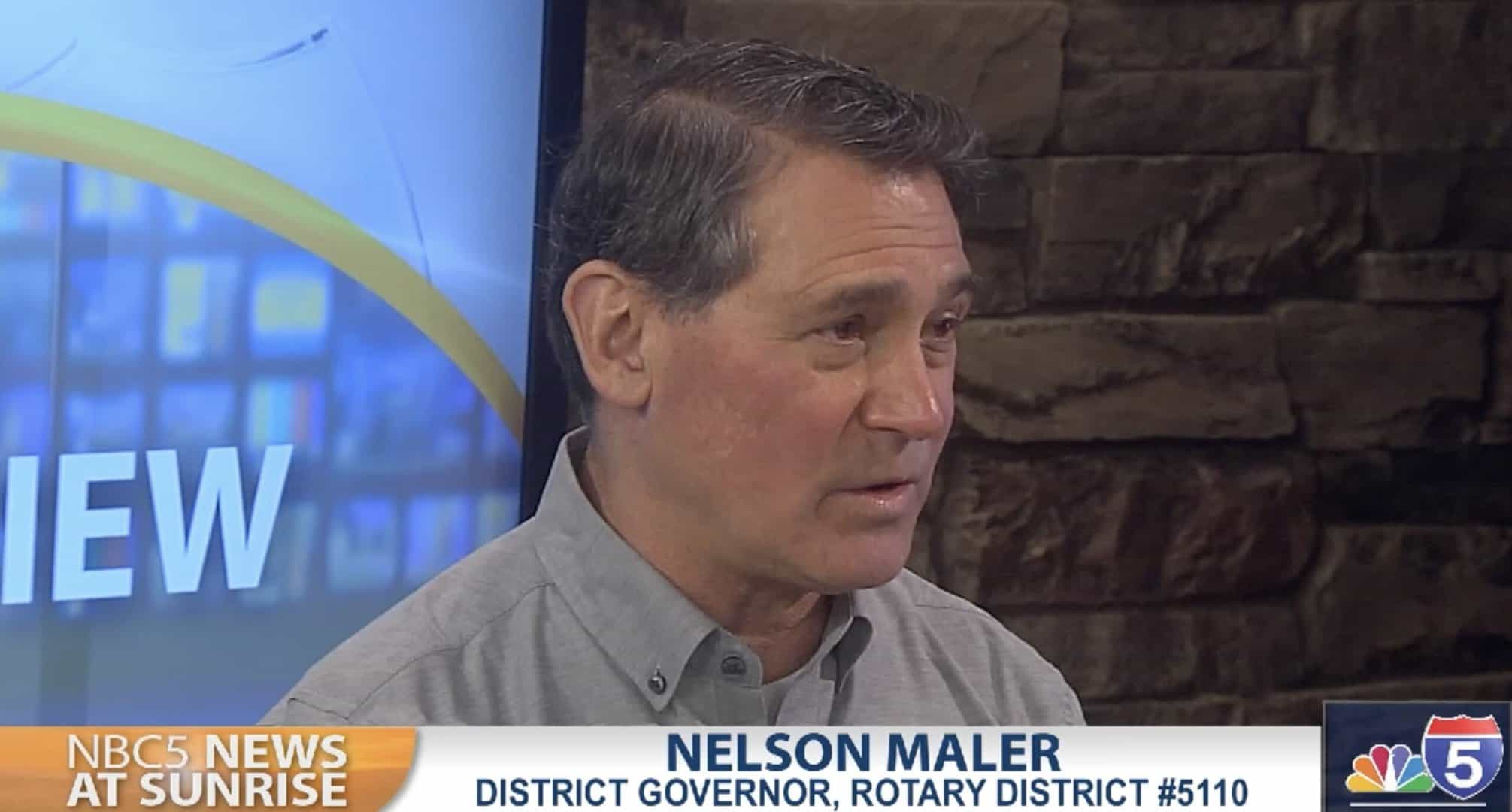 Nelson Maler sits in a news studio for an interview. He is in front of a large projection screen and is wearing a button-up polo shirt with Rotary District 5110 embroidered above the pocket.