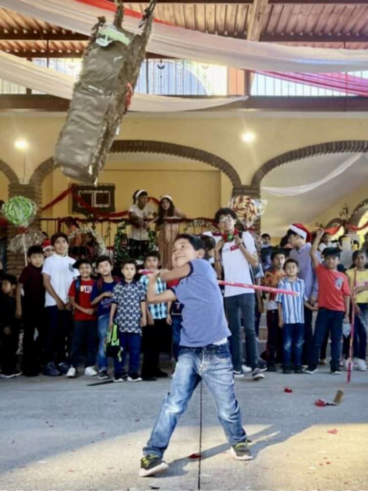 A primary school boy is wearing a blue polo shirt and jeans. He is swinging a pink wooden stick toward a donkey-shaped piñata. He is smiling at an outdoor community center with children lined up behind him watching and waiting for their turns.