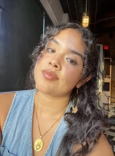 Sophia Olivares has black curly hair. She is wearing a light blue v-neck top with a gold necklace and gold pendant.