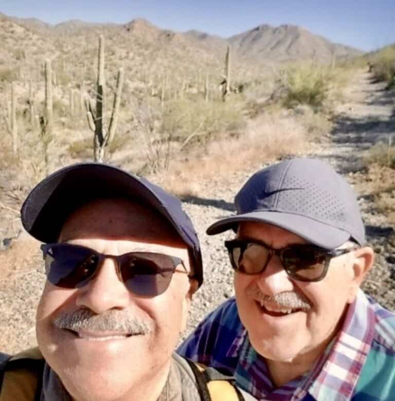 Grandpa Bobby and Grandpa Scott smile in the desert. They are standing in the sun with sunglasses and ballcaps on.