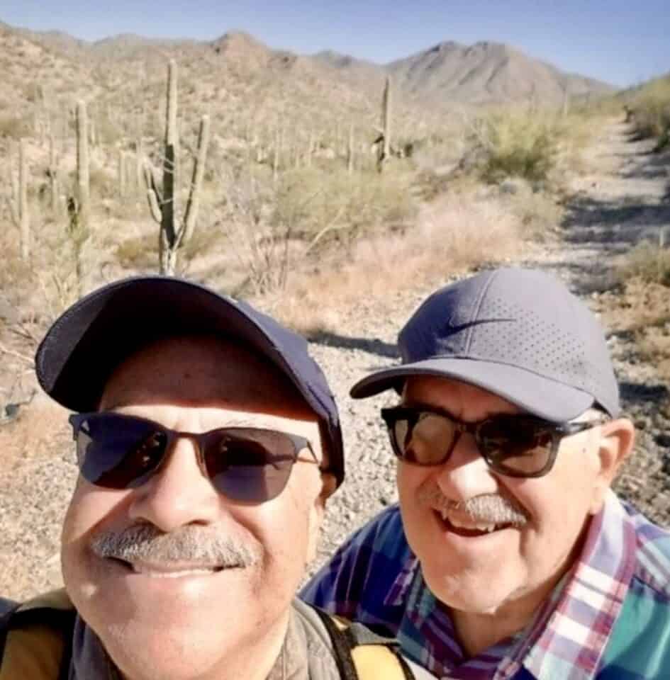 Grandpa Bobby and Grandpa Scott smile in the desert. They are standing in the sun with sunglasses and ballcaps on.