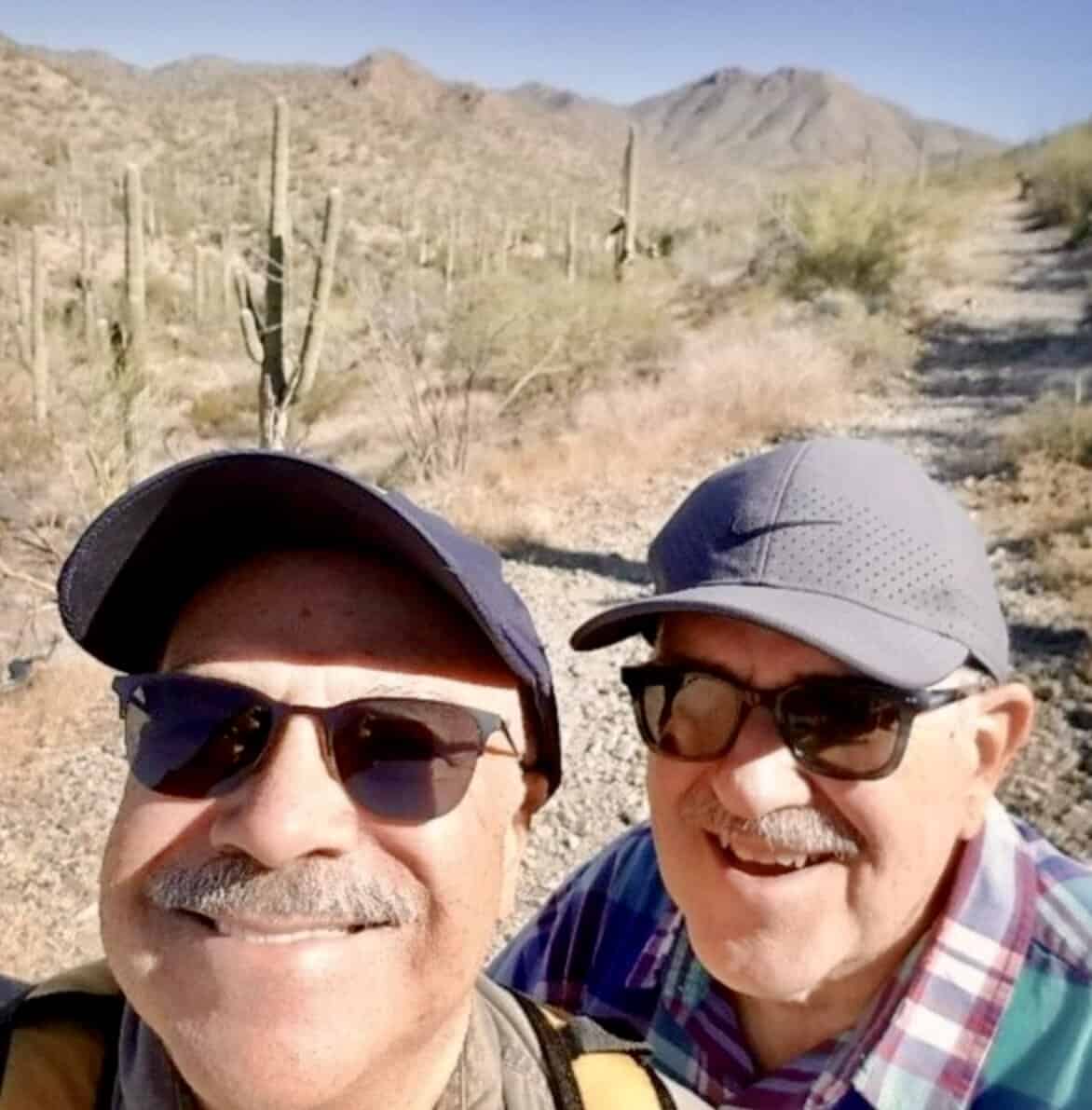 Grandpa Bobby and Grandpa Scott smile in the desert. They are standing in the sun with sunglasses and ballcaps on.
