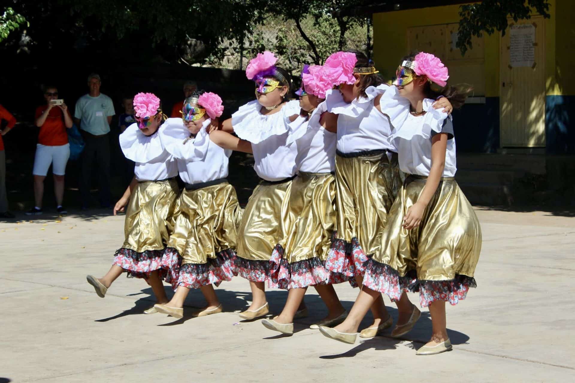 Six primary school girls dance in matching gold skirts with floral trim, white blouses, colorful eye masks, and large pink flowers in their hair. They have their arms around each other and are kicking one foot out in unison.