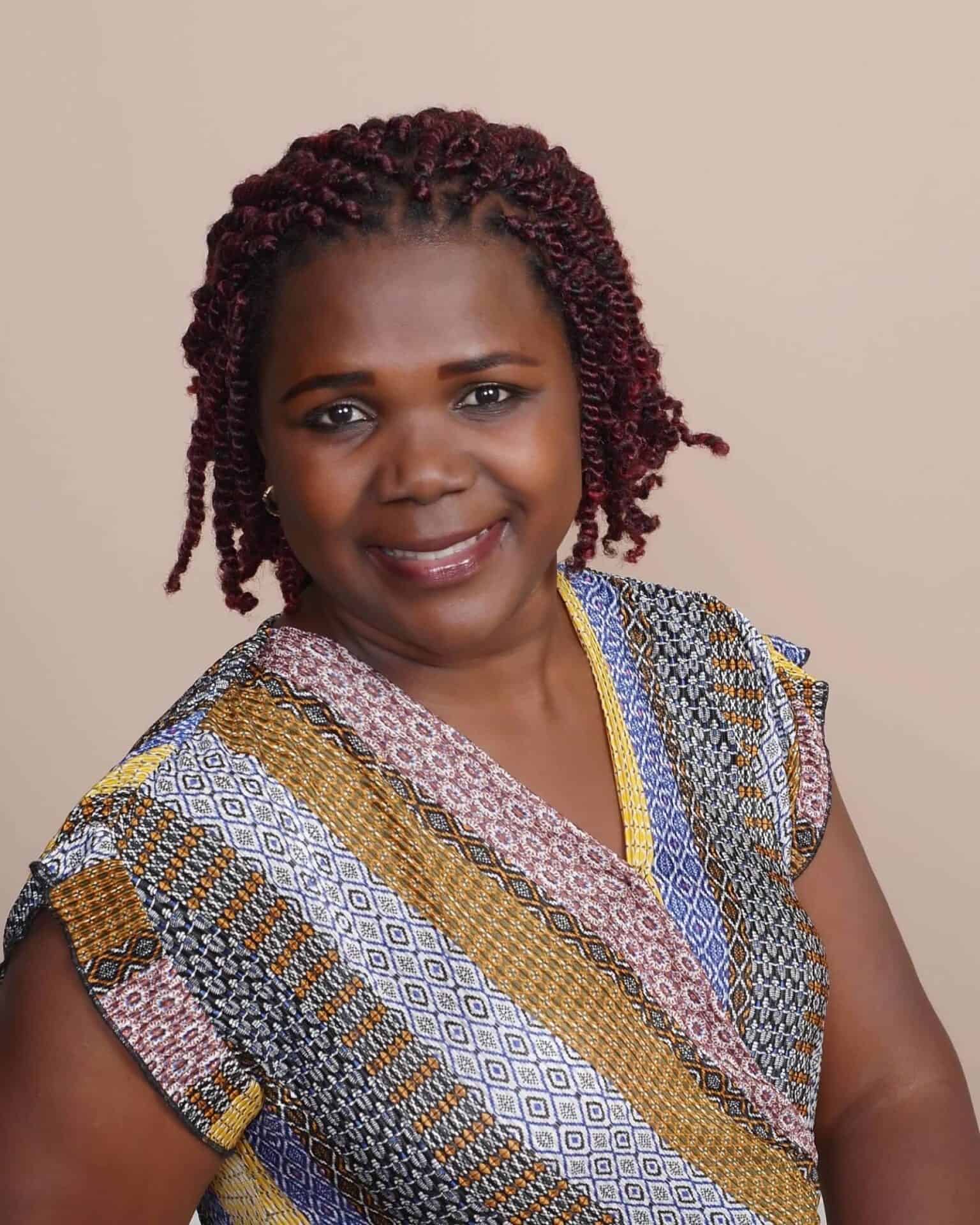 Sylvia Ryan is a Ugandan woman with chin-length hair. She is leaning to her right and smiling at the camera. she is wearing a multi-patterned v-neck short-sleeved shirt.