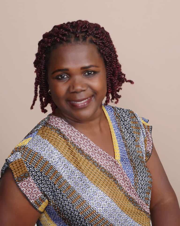Calling All World Travelers 2 Sylvia Ryan is a Ugandan woman with chin-length hair. She is leaning to her right and smiling at the camera. she is wearing a multi-patterned v-neck short-sleeved shirt.