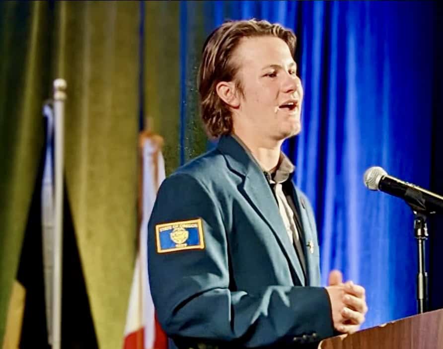 An Rotary Youth Exchange student stands at a podium in front of various flags speaking at a conference. He is wearing a blue blazer and resting his hands below the microphone.