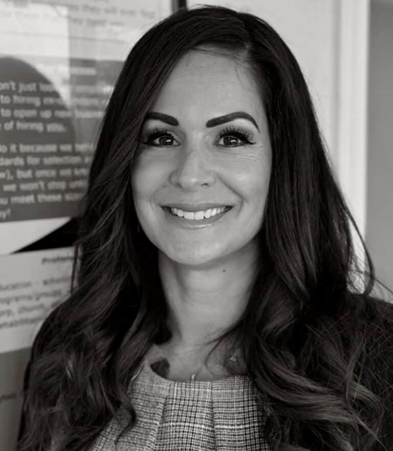 Nancy Pance has long dark hair. She is wearing a plaid top pleated at the neck. She is smiling in a black and white photo.