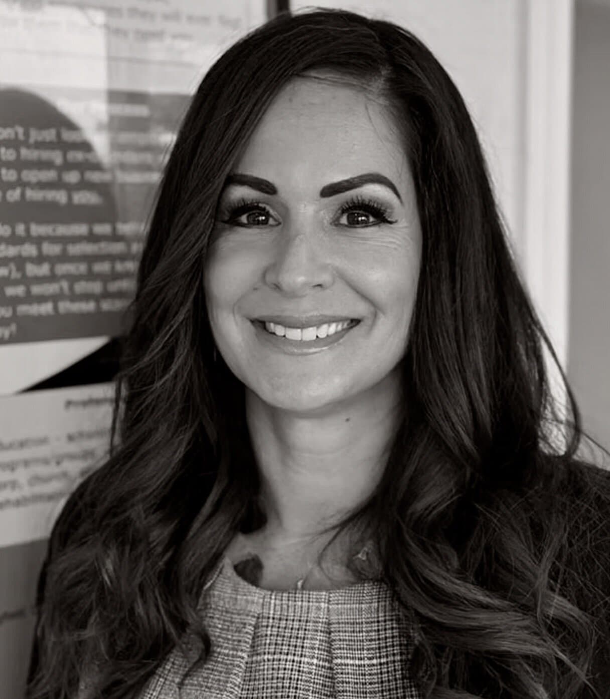 Nancy Pance has long dark hair. She is wearing a plaid top pleated at the neck. She is smiling in a black and white photo.