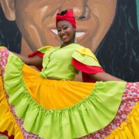 An Afro-Ecuadorian dance artist poses in front of a mural. She is smiling, wearing a traditional marimba dance costume with layered sleeves and a layered skirt in lime green, light yellow, bright red, and a red head wrap.