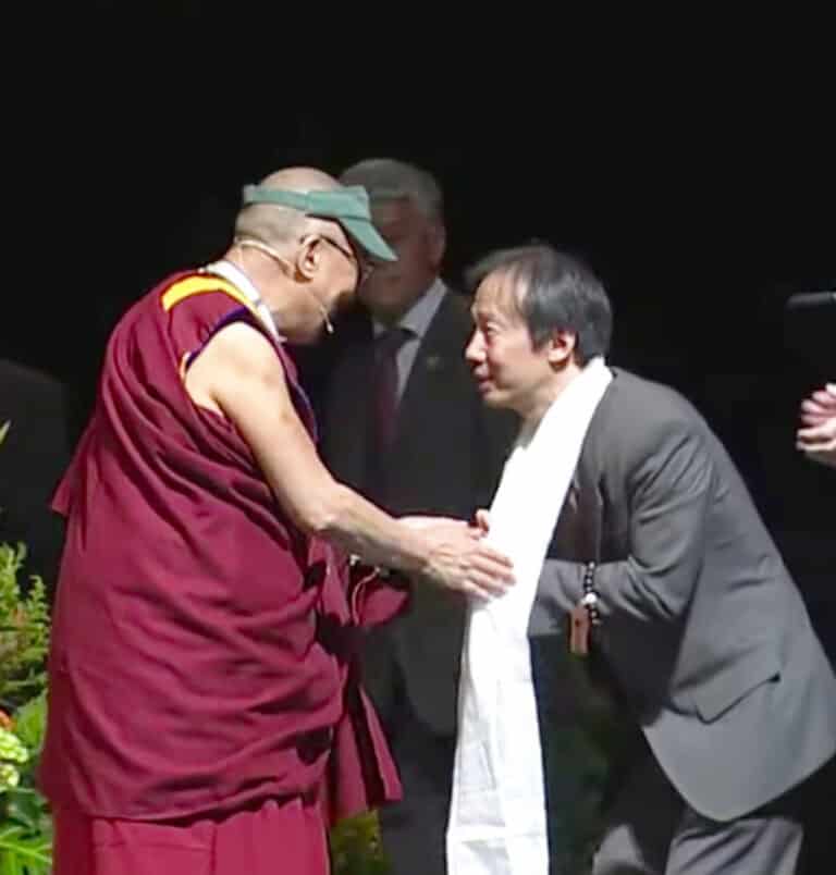 Dr. Mark Unno thanks the 14th Dalai Lama. He is wearing a grey suit and a long white scarf. The Dalai Lama is wearing traditional Buddhist monk vestments and a green UO visor gifted to him. They are clasping hands and smiling at each other.