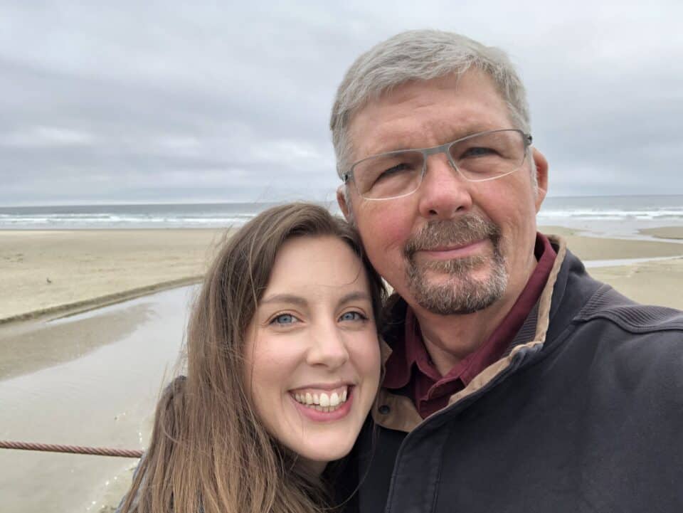 Bruce Allen at the coast with his daughter Rachel. They are smiling at the camera and standing in front of the ocean on an overcast day.