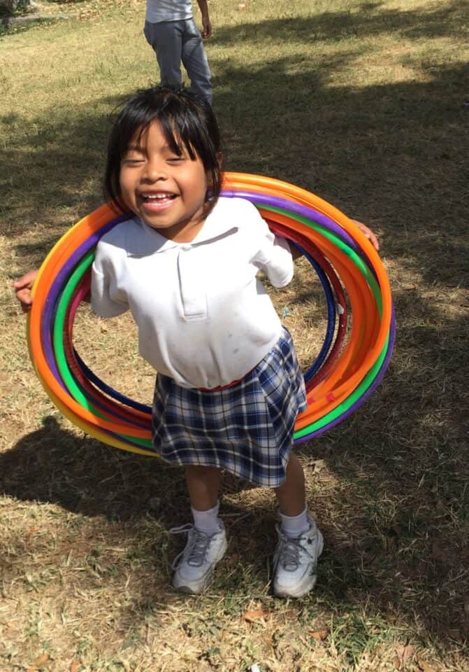 A primary school girl wearing a white blouse and checkered skirt scool uniform smiles at a park. She is "wearing" a small stack of hula hoops.