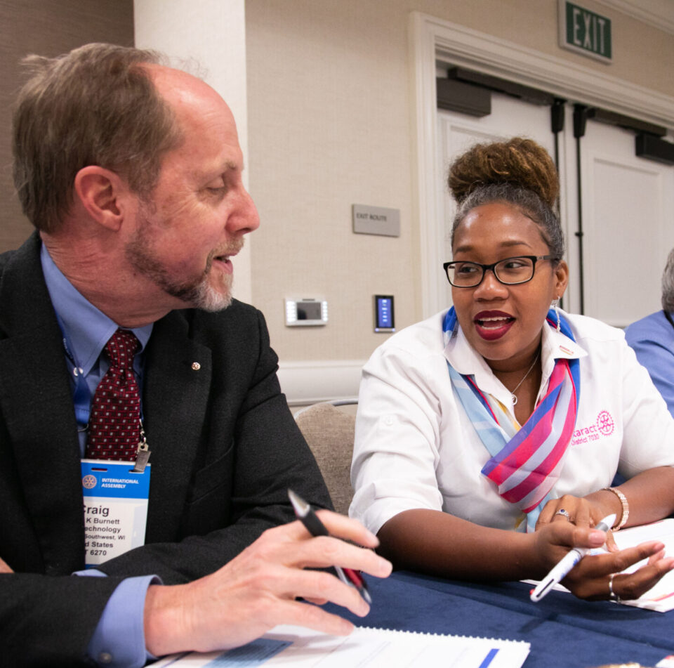 Photo of participants of a Rotary meeting. A man and a woman sit at a conference table discussing ideas.