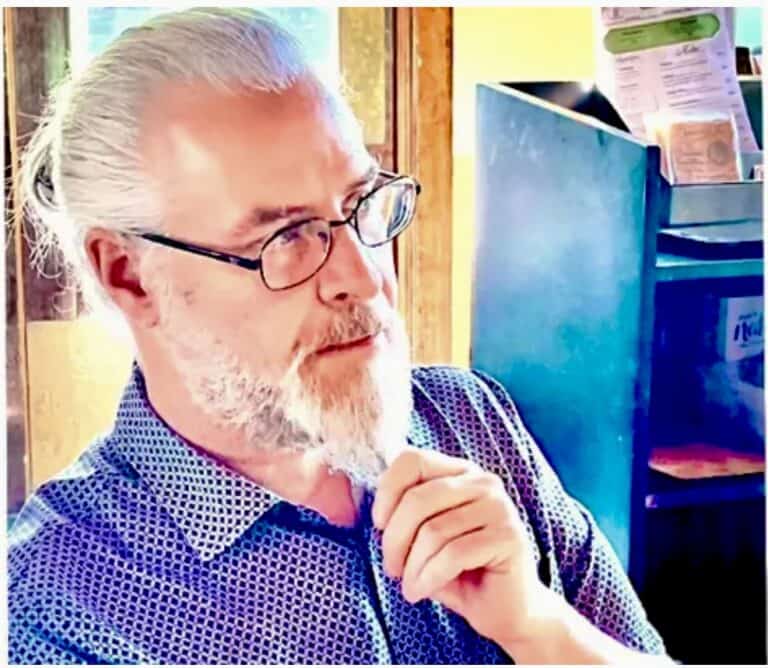 Tom Kamis, owner of the Davis Restaurant looks off-camera. He has gray hair in a ponytail, a gray beard, and glasses. He is wearing a periwinkle-colored polo shirt.