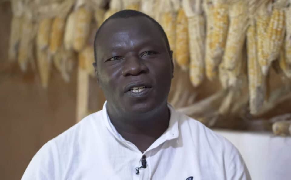 Daniel Wanjama is interviewed on camera. He is wearing a white polo shirt with yellow corn hanging to dry behind him.