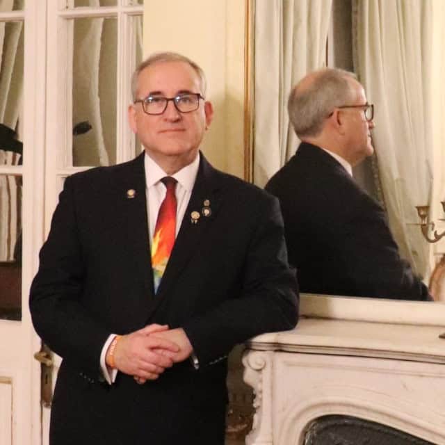 PDG Roberto Silva Perez leans against a marble fireplace mantle. He is wearing a black blazer and multi-colored Rotary necktie with various Rotary pins on his lapel.